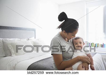 Stock Photo - Mother holding baby son in lap on bed. Fotosearch