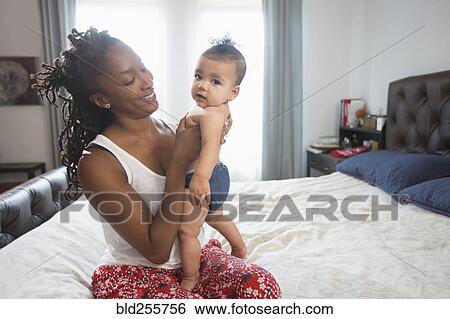 Mother sitting on bed holding baby daughter View Large Photo Image Stock Photograph - Mother sitting on bed holding baby daughter. Fotosearch