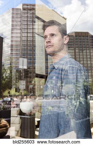 Stock Image - Pensive Caucasian man drinking coffee near window in city. Fotosearch