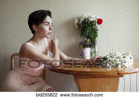 Stock Image - Pensive Caucasian woman sitting at table with flowers. Fotosearch