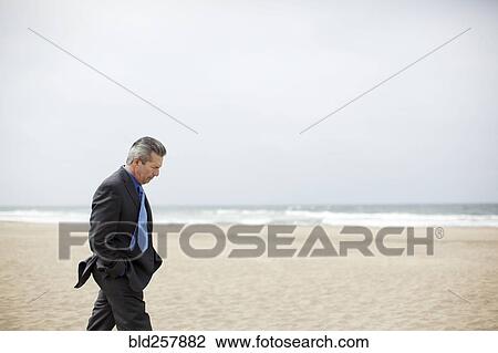 Stock Image - Pensive Hispanic businessman walking on beach. Fotosearch