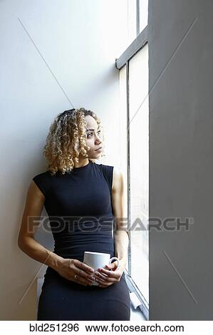 Stock Photograph - Pensive mixed race woman sitting on windowsill drinking coffee. Fotosearch