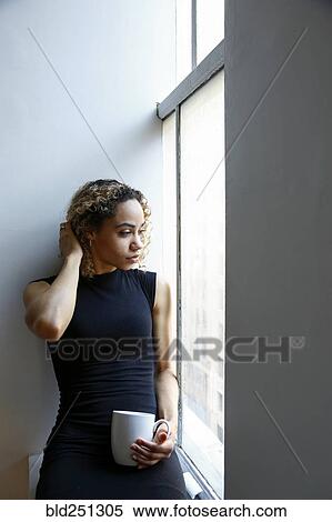 Stock Photography - Pensive mixed race woman sitting on windowsill drinking coffee. Fotosearch