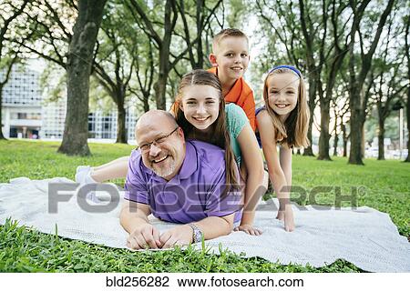Stock Image - Portrait of Caucasian boy and girls laying on back of father in park. Fotosearch