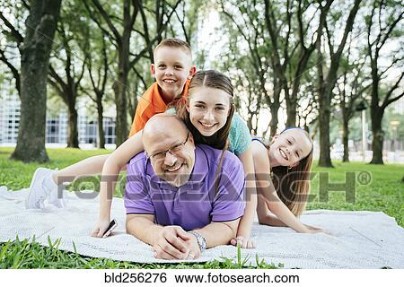 Stock Photograph - Portrait of Caucasian boy and girls laying on back of father in park. Fotosearch