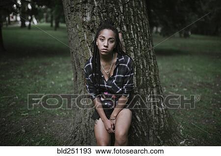 Stock Image - Portrait of serious African American woman leaning on tree. Fotosearch