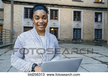 Stock Image - Portrait of smiling Black woman using laptop outdoors. Fotosearch