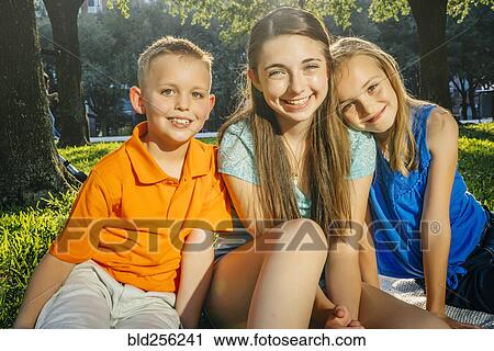 Stock Image - Portrait of smiling Caucasian brother and sisters in park. Fotosearch