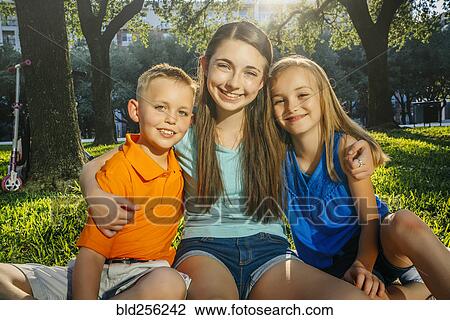 Stock Image - Portrait of smiling Caucasian brother and sisters hugging in park. Fotosearch