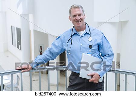 Stock Image - Portrait of smiling Caucasian doctor leaning on railing holding laptop. Fotosearch