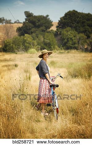 Stock Image - Portrait of smiling Caucasian woman standing in field with bicycle. Fotosearch
