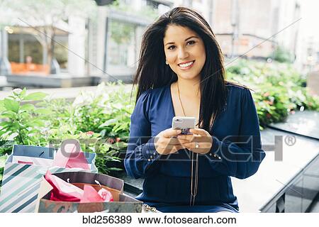 Stock Photo - Portrait of smiling Hispanic woman with shopping bags texting on cell phone. Fotosearch