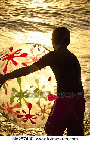 Stock Image - Silhouette of Caucasian boy holding beach ball at sunset. Fotosearch