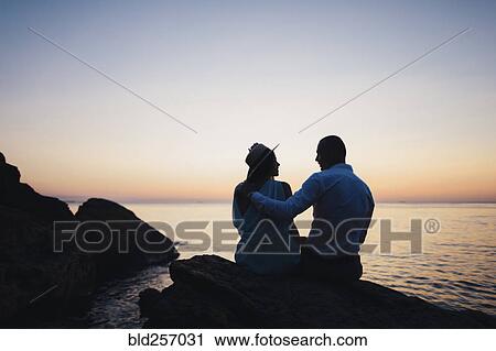 Stock Image - Silhouette of Caucasian couple sitting on rock near ocean at sunset. Fotosearch