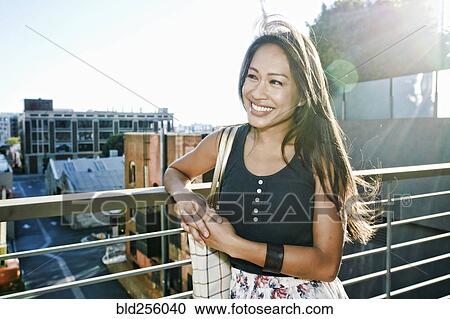 Stock Image - Smiling Asian woman leaning on rooftop. Fotosearch