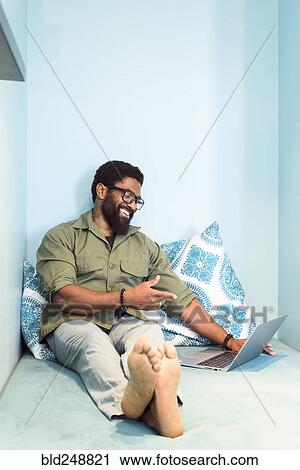Stock Image - Smiling black man sitting on bed pointing at laptop. Fotosearch