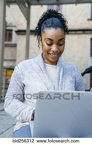 Stock Image - Smiling Black woman using laptop outdoors. Fotosearch