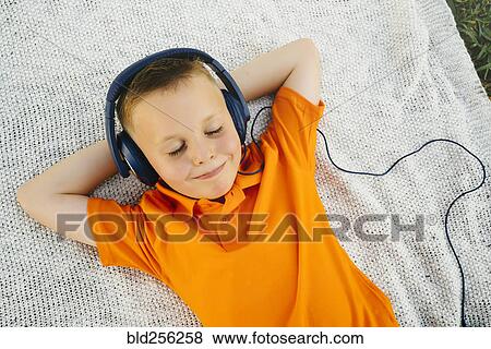 Stock Photo - Smiling Caucasian boy laying on blanket in park listening to headphones. Fotosearch