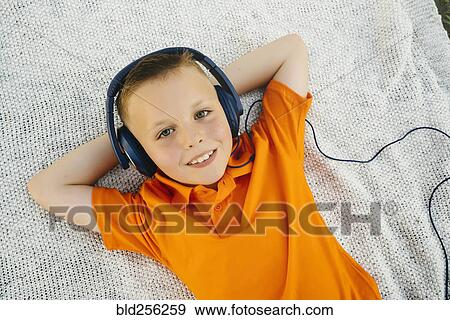 Stock Photo - Smiling Caucasian boy laying on blanket in park listening to headphones. Fotosearch
