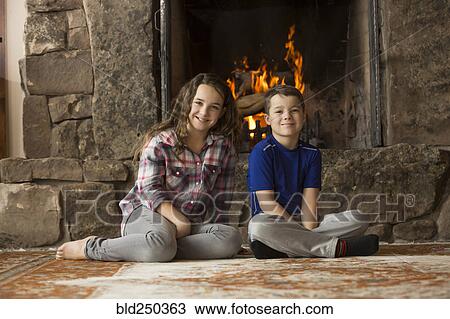 Stock Image - Smiling Caucasian brother and sister sitting on floor near fireplace. Fotosearch