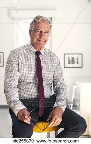 Stock Photo - Smiling Caucasian businessman sitting on stool holding eyeglasses. Fotosearch