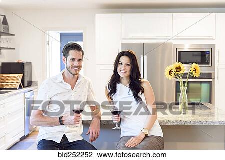 Stock Image - Smiling Caucasian couple drinking red wine in kitchen. Fotosearch