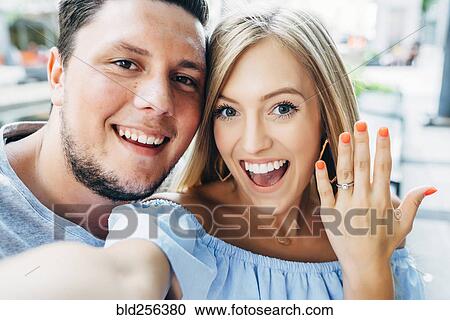 Stock Image - Smiling Caucasian couple posing for selfie with engagement ring. Fotosearch