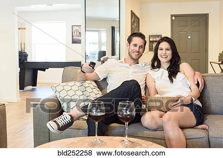 Stock Photo - Smiling Caucasian couple sitting on sofa watching television. Fotosearch