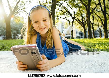 Stock Image - Smiling Caucasian girl laying on blanket in park reading digital tablet. Fotosearch