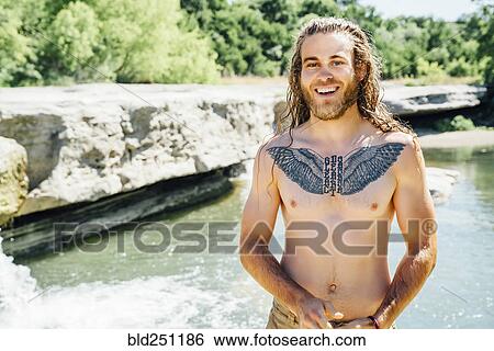 Stock Photograph - Smiling Caucasian man with chest tattoo standing near river. Fotosearch
