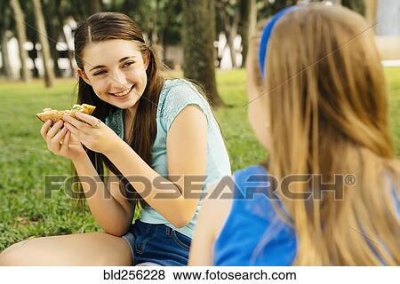 Stock Photo - Smiling Caucasian sisters eating food at picnic. Fotosearch