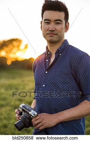 Stock Photo - Smiling Chinese man holding camera in field. Fotosearch
