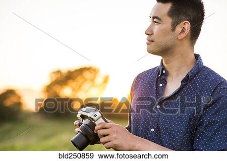 Smiling Chinese man holding camera in field View Large Photo Image Stock Photo - Smiling Chinese man holding camera in field. Fotosearch