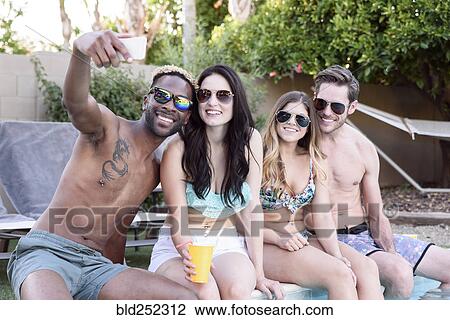 Stock Image - Smiling friends sitting poolside posing for cell phone selfie. Fotosearch