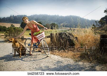 Stock Image - Smiling Hispanic woman standing with bicycle petting dog. Fotosearch