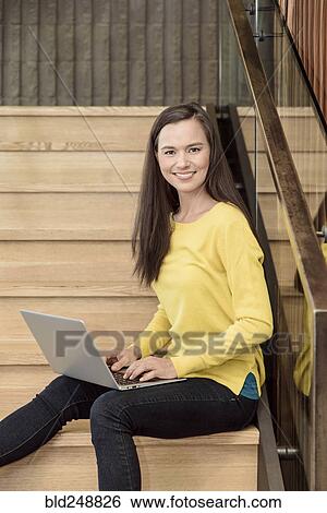 Stock Photograph - Smiling mixed race woman sitting on staircase using laptop. Fotosearch