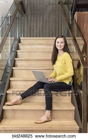 Stock Photography - Smiling mixed race woman sitting on staircase using laptop. Fotosearch