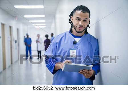 Stock Image - Smiling nurse leaning on wall in hospital using digital tablet. Fotosearch