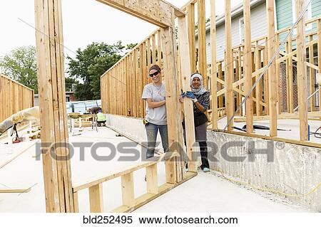 Stock Photography - Smiling volunteers holding framed wall at construction site. Fotosearch