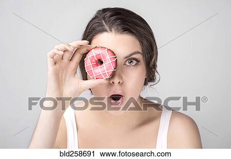 Stock Image - Sur1ised Caucasian woman holding donut over eye. Fotosearch