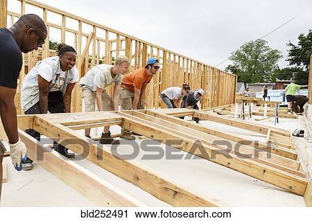 Stock Image - Volunteers lifting framed wall at construction site. Fotosearch