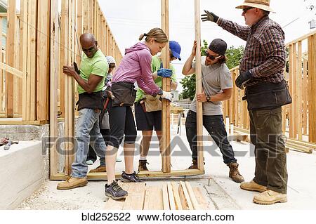 Stock Image - Volunteers lifting framed wall at construction site. Fotosearch