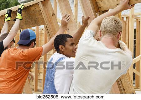 Stock Photograph - Volunteers lifting framed wall at construction site. Fotosearch