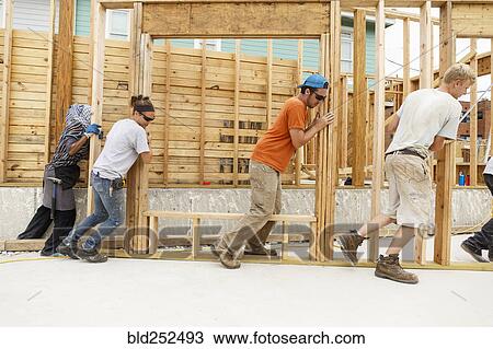 Stock Image - Volunteers pushing framed wall at construction site. Fotosearch