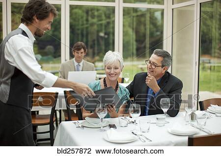 Stock Image - Waiter 1esenting menu to couple in restaurant. Fotosearch