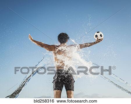 Stock Photograph - Water splashing on back of Hispanic man holding soccer ball. Fotosearch