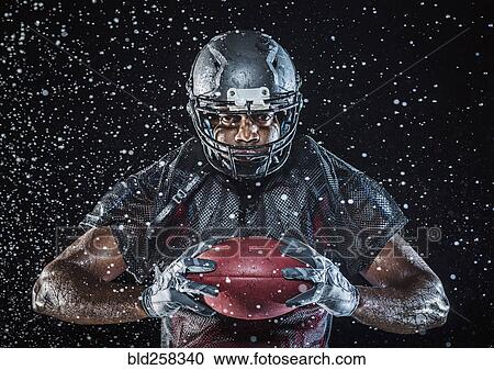 Stock Image - Water splashing on Black football player holding football. Fotosearch
