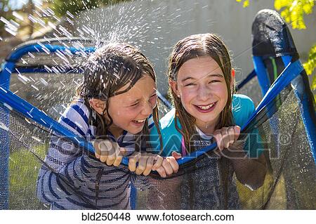 Stock Photo - Water splashing on Caucasian girls leaning on netting. Fotosearch