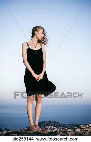 Picture - Wind blowing dress of Caucasian woman standing on rock near ocean. Fotosearch