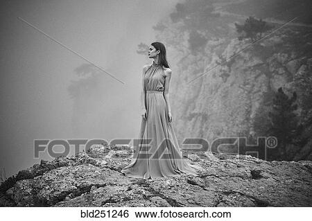 Stock Photograph - Woman wearing dress standing on rock in fog. Fotosearch